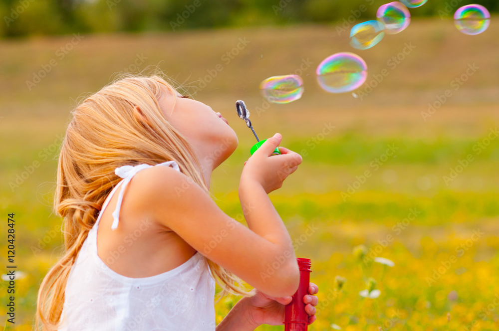 Little girl blowing soap balloons while sitting on spring meadow  - obrazy, fototapety, plakaty 