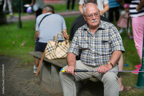 Senior beim Minigolf. Er sitzt auf der Bank, wartet auf seinen Einsatz und beobachtet die anderen Spieler.