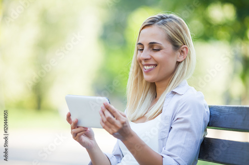 Young happy woman is sitting in park and using digital tablet. 
