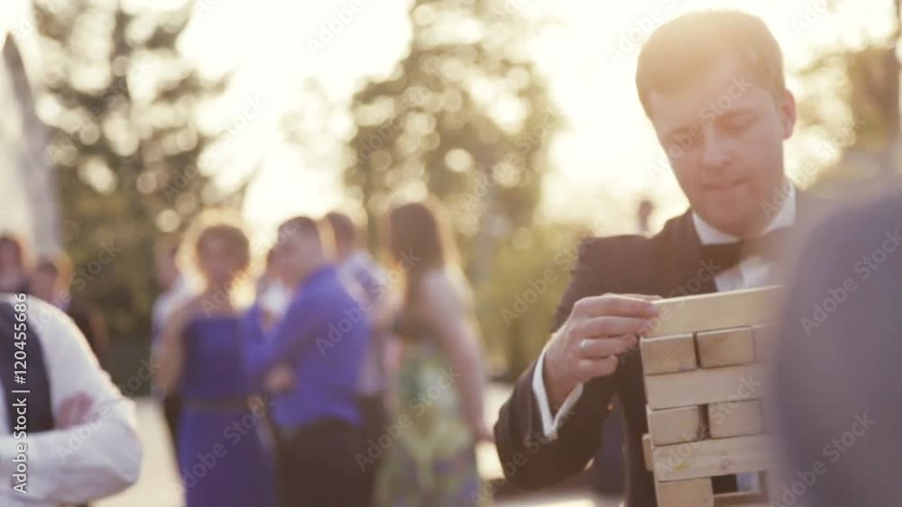 Wedding celebration, a bridegroom in an elegant black wedding costume, white shirt and bow tie playing jenga with his friends during wedding party. Outside shooting, sunset, trees. Wedding atmosphere.