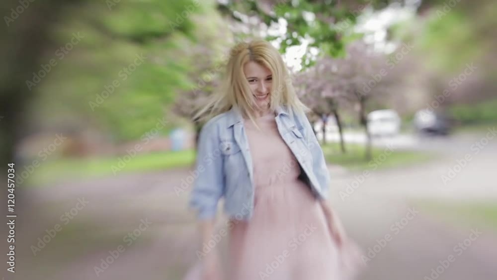 Young blonde girl in pink dress and jeans jacket running down the blossoming alley, turns o camera and smiles. Female portrait. Outside shooting, spring time. Blossoming cherry trees . Lensbaby. Young blonde girl in pink dress and jeans jacket running down the blossoming alley, turns o camera and smiles. Female portrait. Outside shooting, spring time. Blossoming cherry trees . Lensbaby.
