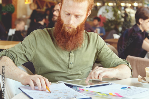 Serious young redhead Caucasian businessman dressed casually holding touch pad pc, looking at screen with concentrated expression while filling in some papers, working at cafeteria during lunch break