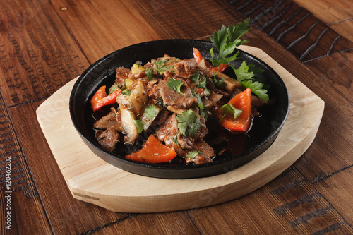 Fried lamb meat slices with vegetables in a pan. Wooden background.