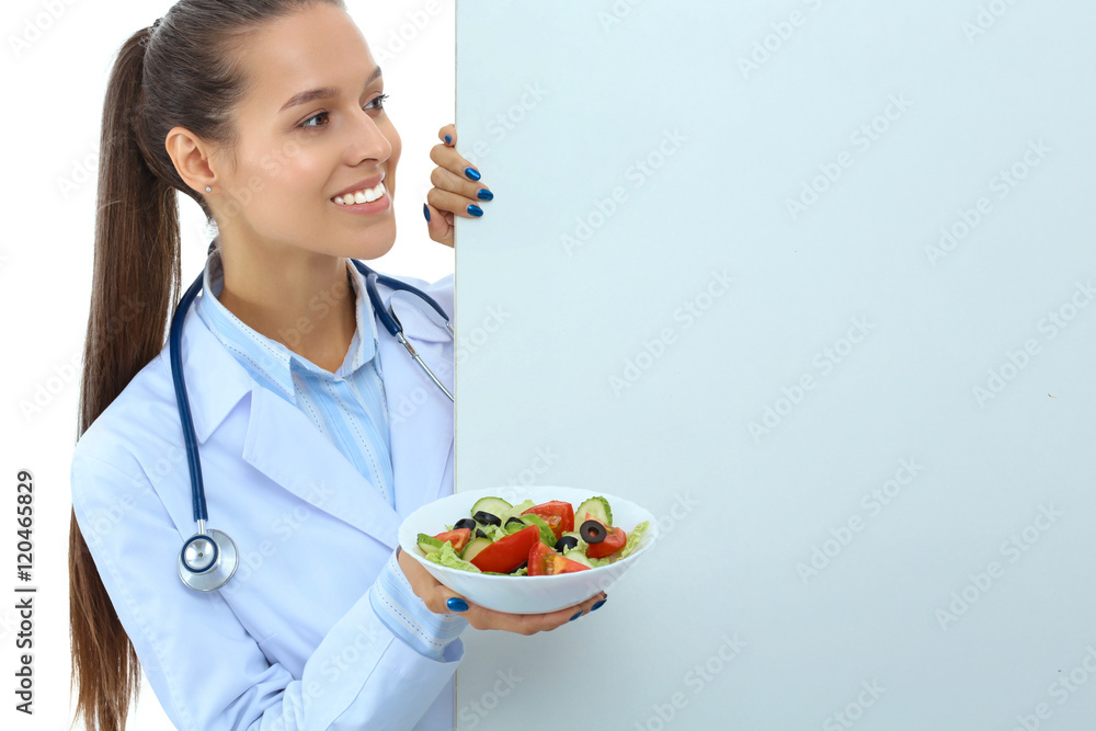 Portrait of a beautiful woman doctor holding a plate with fresh vegetables standing near blank Portrait of a beautiful woman doctor holding a plate with fresh vegetables standing near blank