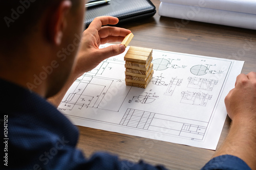 The man (businessman, architect) draws a plan, graph, design on large sheet of paper at office desk and builds model house from wooden blocks 
