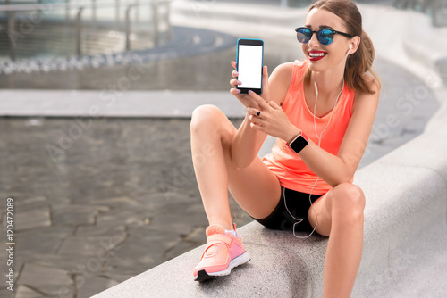 Young sports woman showing phone with white screen to copy paste sitting on the modern bench in the city photo