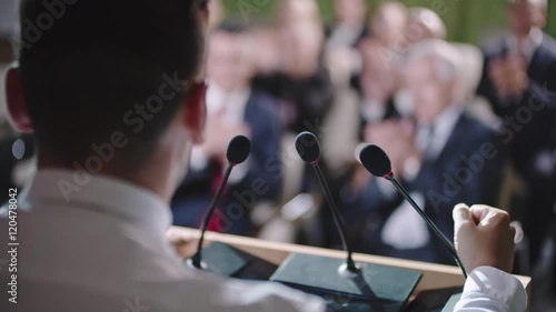 Over the shoulder shot of politician making speech behind tribune before applauding audience on political rally 