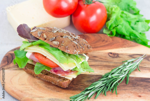 Sandwich with ham, tomato, cheese and lettuche on cutting board with rosemary on it. Closeup fast morning breakfast. photo