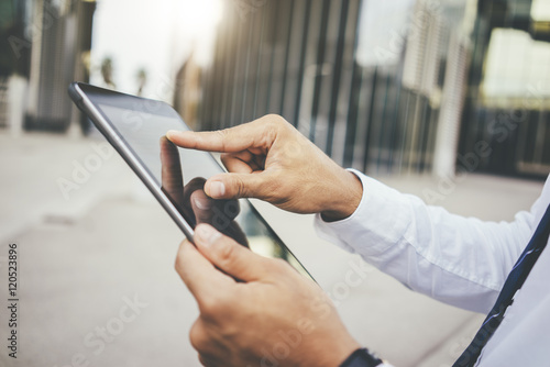 Close-up of male hands typing text message on digital tablet, businessman holding and using modern tablet pc outside, focus on hands, office buildings in the background