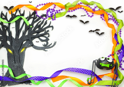 Halloween border of a rough textured wooden cutout of bare tree shape painted black. A silly spider  bats and purple  green and orange ribbons and beads frame the copy space on white background