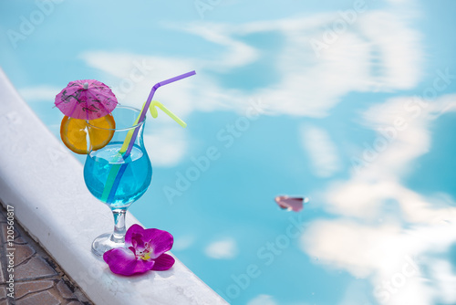 Fototapeta Naklejka Na Ścianę i Meble -  Blue cocktail decorated slice of orange and exotic flowers near pool