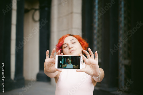 portrait of a girl doing selfie. girl in light pink dress with red dyed hair, pictures of themselves on their mobile camera phone, sitting on the stairs summer day