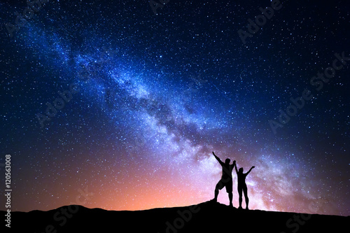Fototapeta Naklejka Na Ścianę i Meble -  Milky Way. Colorful night sky with stars and silhouette of standing happy man and woman with raised  up arms on the hill. Blue milky way with people on the mountain. Background with beautiful universe