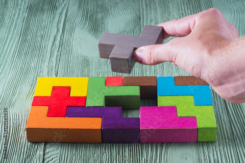 Hand holding wooden puzzle element. Hand sets the last element of the puzzle. The concept of logical thinking. Geometric shapes on a wooden background. Tetris toy wooden blocks.