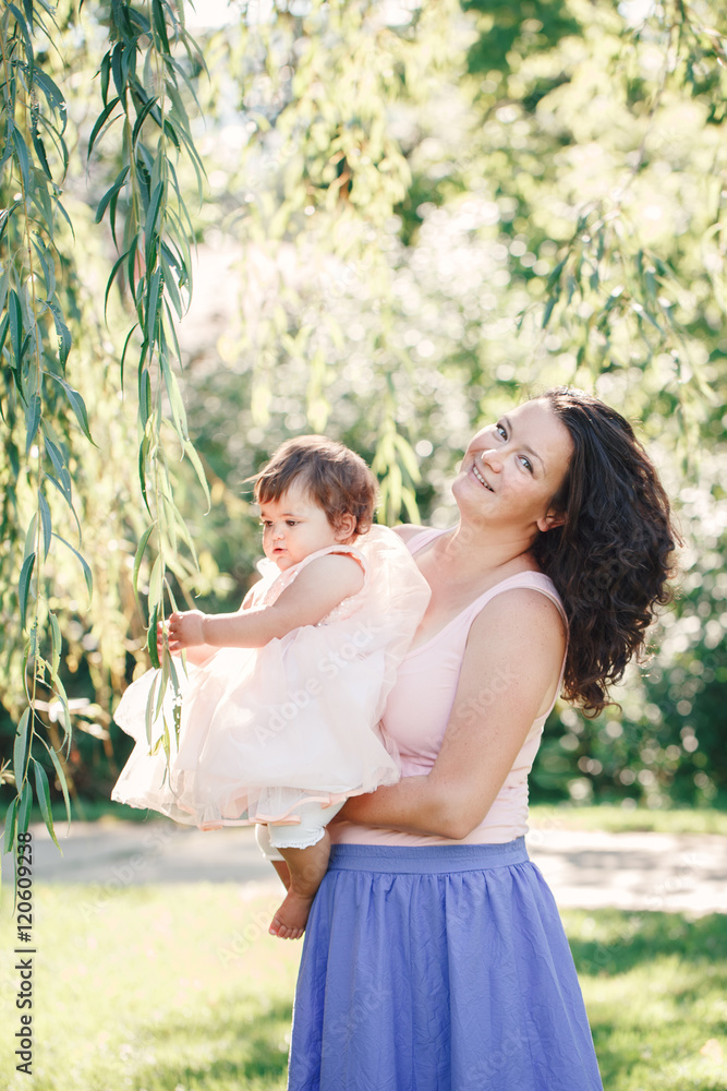 Naklejka premium Lifestyle group portrait of smiling white Caucasian brunette mother holding hugging daughter in pink dress looking in camera on sunny spring summer day in park outside