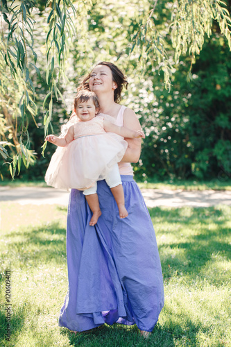 Lifestyle group portrait of smiling white Caucasian brunette mother holding hugging daughter in pink dress dancing with baby child on sunny summer day in park