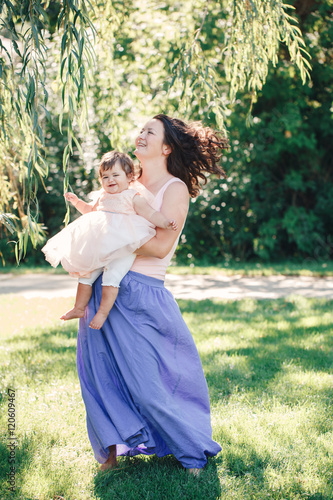 Lifestyle group portrait of smiling white Caucasian brunette mother holding hugging daughter in pink dress dancing with baby child on sunny summer day in park