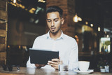 Young hipster guy sitting at modern cafe and using digital tablet, confident male hispanic businessman reading information at internet via tablet pc during a break, office worker, visual effects