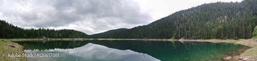 Panoramic image of lake landscape in the mountains, with pine trees reflecting on the water, on a cloudy day; nature with no people. Black lake, Durmitor, Montenegro. Horizontal panorama.