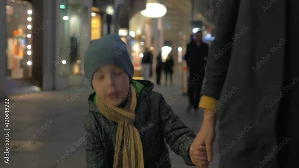 Little boy in overclothes holding mothers hand and talking to her while they walking in city street. Evening outing in pedestrian shopping area Little boy in overclothes holding mothers hand and talking to her while they walking in city street. Evening outing in pedestrian shopping area