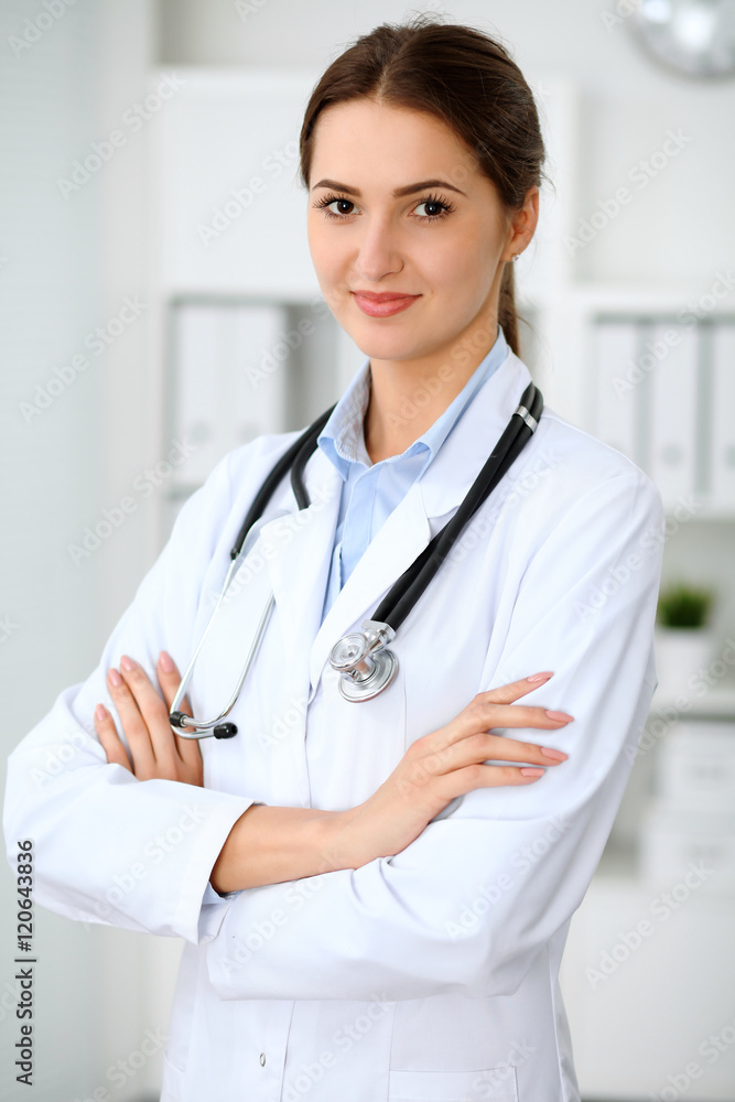 Young brunette female doctor standing with arms crossed and smiling at hospital. Health care, insurance and help concept. Physician ready to examine patient Young brunette female doctor standing with arms crossed and smiling at hospital. Health care, insurance and help concept. Physician ready to examine patient
