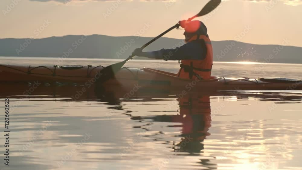 Slow motion tracking of man with beard paddling kayak on lake at sunset, chain of mountains visible in distance Slow motion tracking of man with beard paddling kayak on lake at sunset, chain of mountains visible in distance