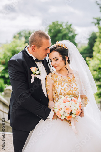 Beautiful brunette bride in elegant dress and handsome groom in blue suit at wedding day lovely hugging outdoors on nature. Happy newlywed woman and man embracing in green park. Loving wedding couple. photo