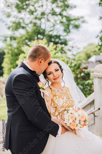 Beautiful brunette bride in elegant dress and handsome groom in blue suit at wedding day lovely hugging outdoors on nature. Happy newlywed woman and man embracing in green park. Loving wedding couple. photo