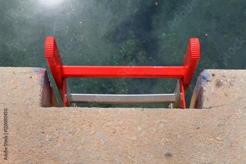 Bright red metal ladder on a concrete quayside leading down to the clear green water of a harbour, with the sun reflecting