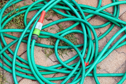 Green garden water hose with red and white nozzle coiled up untidily on a stone block background with weeds. photo