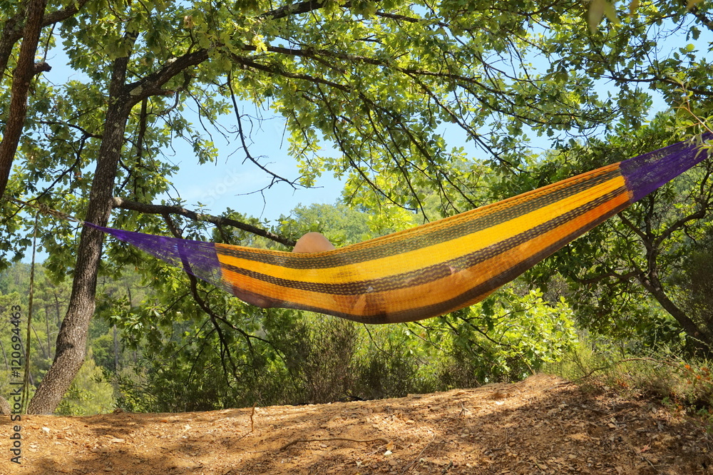 Person resting in hammock with yellow stripes hung between sun dappled trees on a sunny day Person resting in hammock with yellow stripes hung between sun dappled trees on a sunny day