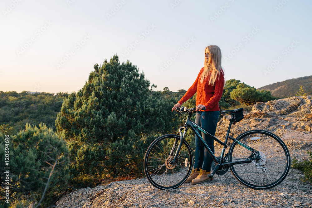 Fototapeta premium attractive young blonde woman in red sweater, blue jeans and yellow boots stands with bike on mountain road and looking forward