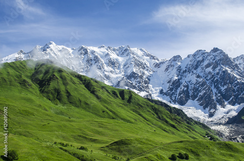 Mountain landscape with emerald slopes on the foreground and steep face of rocks ice and snow on the background in gorgeous summer day