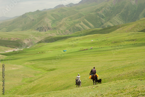 Two riders on horseback go away in the valley between the green mountains. Kyrgyzstan.