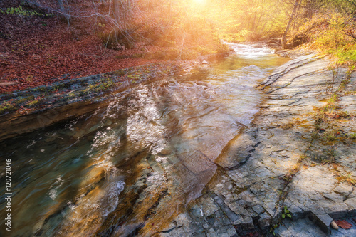 Beautiful fall scene on a clear mountain river with small waterfall and rapids, gravel with with colorful rocky bottom and fallen leaves at sunny day.