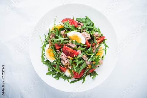 Fresh salad with tuna, tomatoes, eggs, arugula and mustard on white textured background top view. Healthy food.