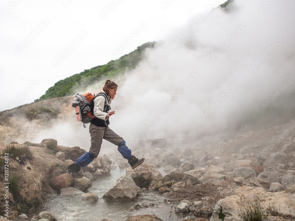 Adult woman with a backpack jumping over a hot stream in a smoking crater of the volcano Mutnovsky on Kamchatka in Russia against the background of a hill with trees and sky with clouds