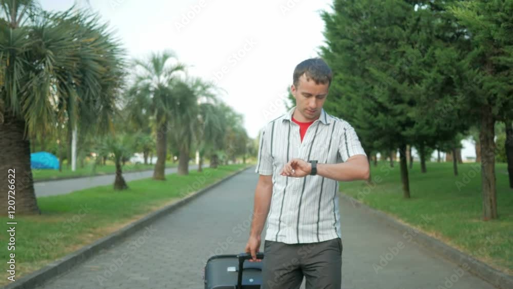 Young man tourist is with a large suitcase on wheels around the city park. Comes message on smart watches, the man stopped and dictates the answer. Young man tourist is with a large suitcase on wheels around the city park. Comes message on smart watches, the man stopped and dictates the answer.