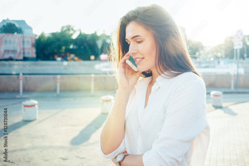 A cute brunette girl in white shirt is talking on a mobile phone while standing on a street. Charming woman with long brown hairs is holding a smartphone while walking down the street on a sunny day.