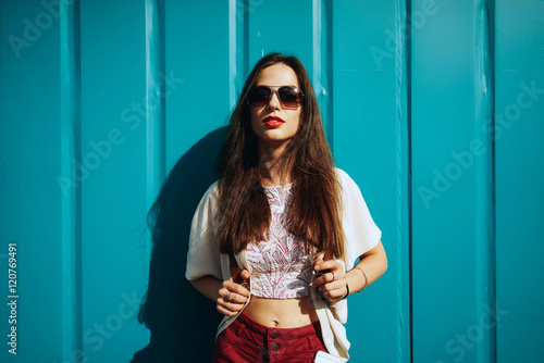 A beautiful model look woman with long brown hair wearing casual clothes is posing on the light blue background. An european brunette female wearing sunglasses is looking at the camera. © JKstock