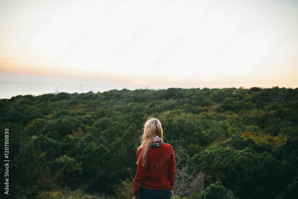 attractive young blonde woman  in red sweater looks forward on the background of forest. female tourist enjoying view. Rear view.