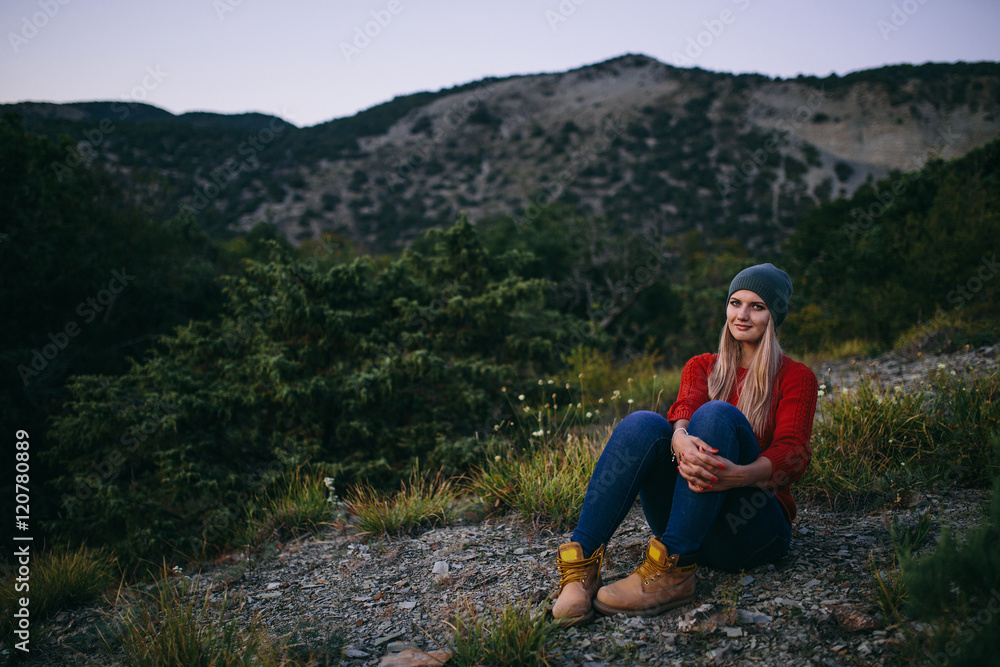 portrait of a beautiful young blonde woman in hat, red sweater, blue jeans and yellow boots sits outdoor on the background of mountain and forest