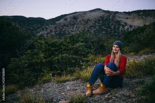 portrait of a beautiful young blonde woman in hat, red sweater, blue jeans and yellow boots sits outdoor on the background of mountain and forest