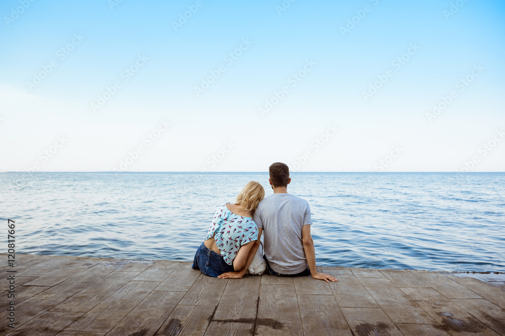 Fototapeta premium Couple sitting back to camera with French bulldog near sea.
