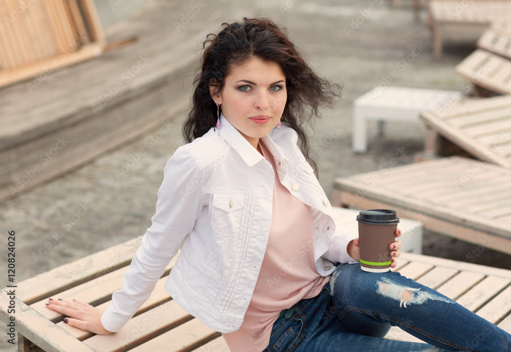 Beautiful tall girl with long hair brunette in jeans standing on old wooden planks with a cup of coffee in hand on a warm summer evening Beautiful tall girl with long hair brunette in jeans standing on old wooden planks with a cup of coffee in hand on a warm summer evening