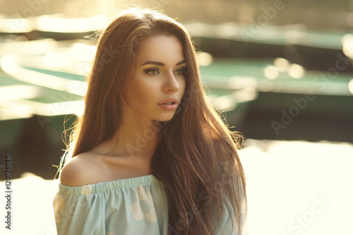 Portrait of young cute woman looking away and posing outdoors in sunset light against bokeh background of boats on water