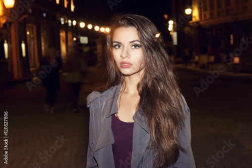 Closeup fashion portrait of young beautiful pretty woman with long curly hair walking in night city and looking at you