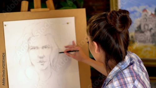Girl artist paints portrait of woman with pencil. 4k. Girl painter seen from behind. Girl paints by pencil on easel.