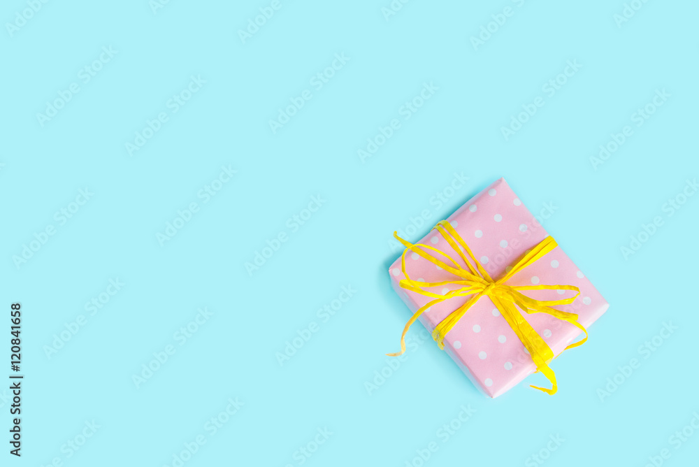 Top view of a gift box wrapped in pink dotted paper and tied yellow bow over light blue background.