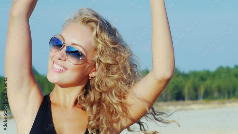 An attractive young woman in sunglasses posing at the camera at the beach. Smiling An attractive young woman in sunglasses posing at the camera at the beach. Smiling